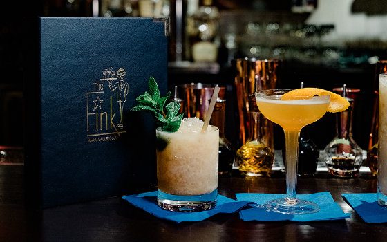 A craft cocktail topped with mint and a coupe cocktail garnished with a peach slice on a bar at The Fink, the speakeasy-style cocktail bar inside Napa River Inn, with The Fink menu propped beside the drinks.