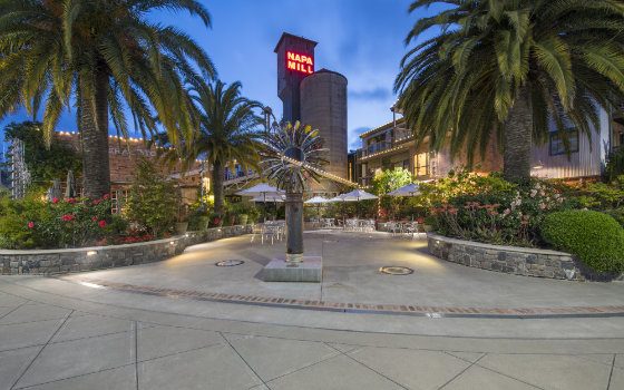 Twilight view of the Historic Napa Mill courtyard at Napa River Inn with palm trees, garden lighting, and the iconic red Napa Mill tower glowing above the river-facing patio.