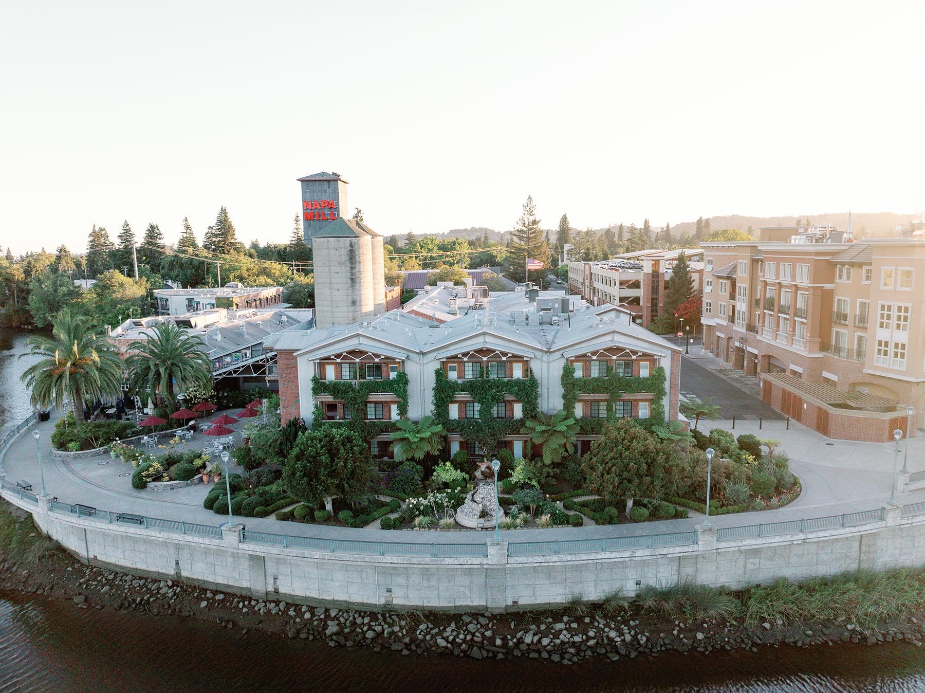 Aerial photo of Napa River Inn and the Historic Napa Mill complex along the Napa River at golden hour, showing the ivy-covered Plaza Building, the iconic red Napa Mill tower, and the surrounding Downtown Napa Riverfront District.