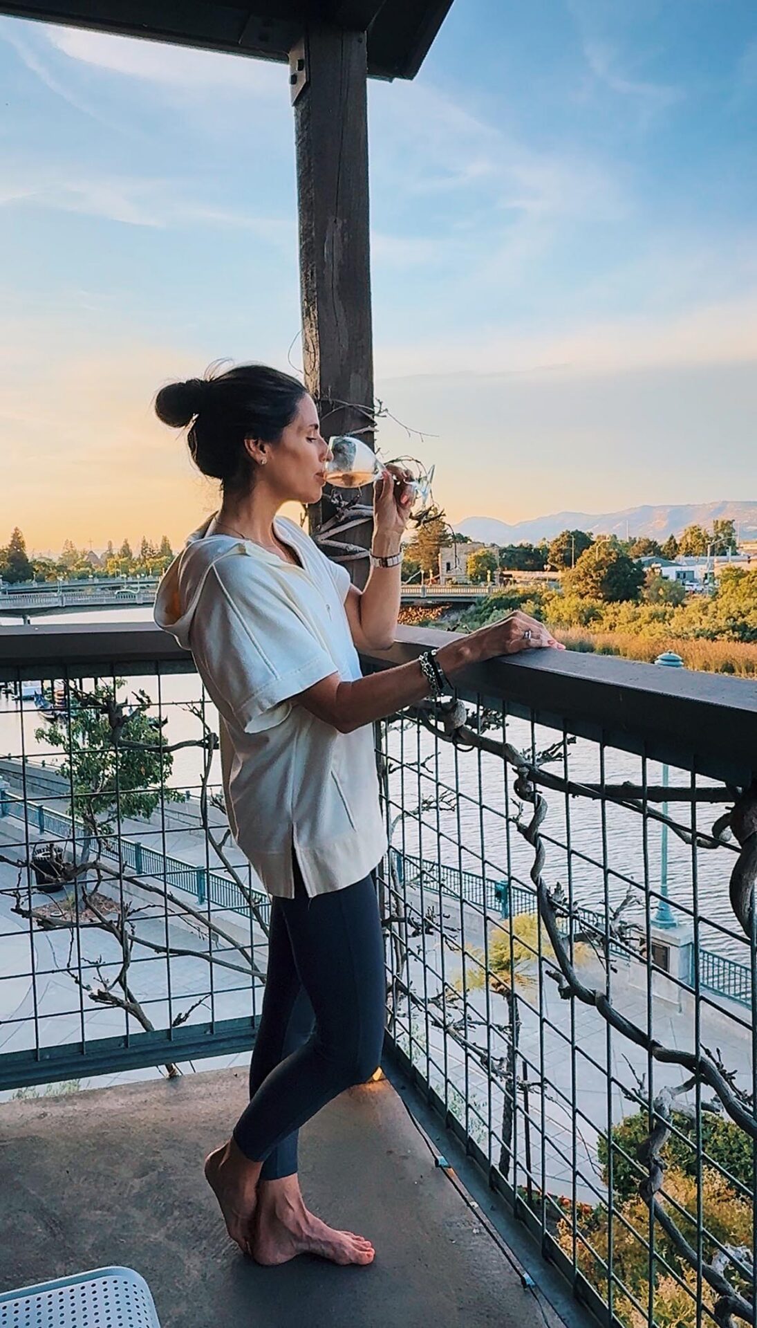 A woman in a soft sweater sips white wine on a private balcony at Napa River Inn overlooking the Napa River and Downtown Napa at sunset.
