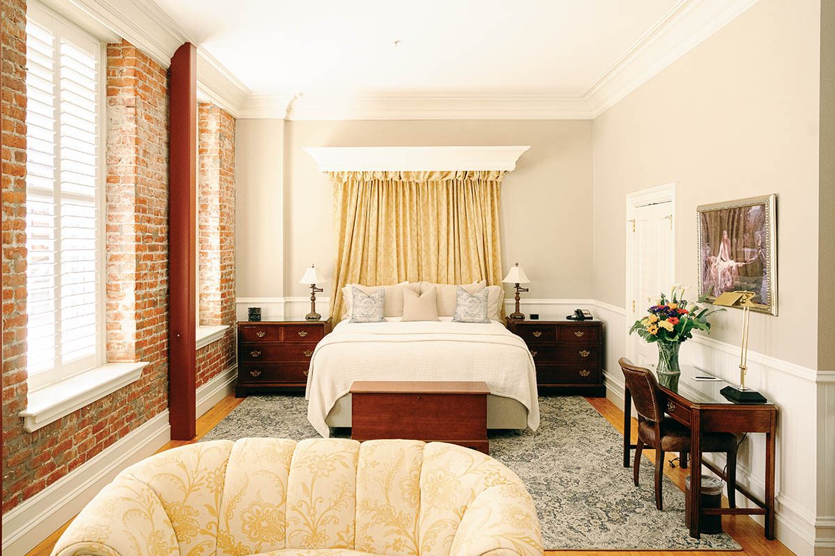 Hatt Suite at Napa River Inn featuring a king bed with cream linens, exposed red brick wall, original wood-shuttered windows, and a yellow draped canopy headboard inside the historic Hatt Building.