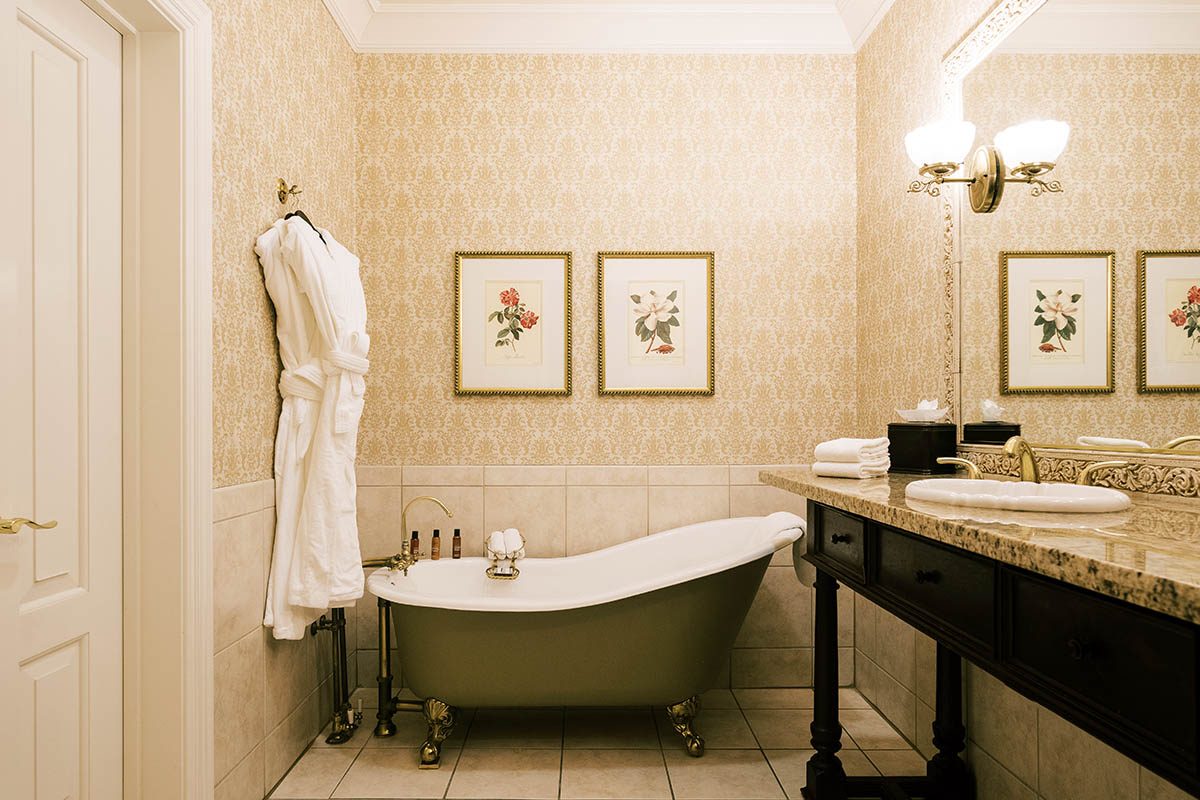 Historic Deluxe bathroom at Napa River Inn with a clawfoot soaking tub, brass fixtures, marble vanity, plush white robes, and damask wallpaper.