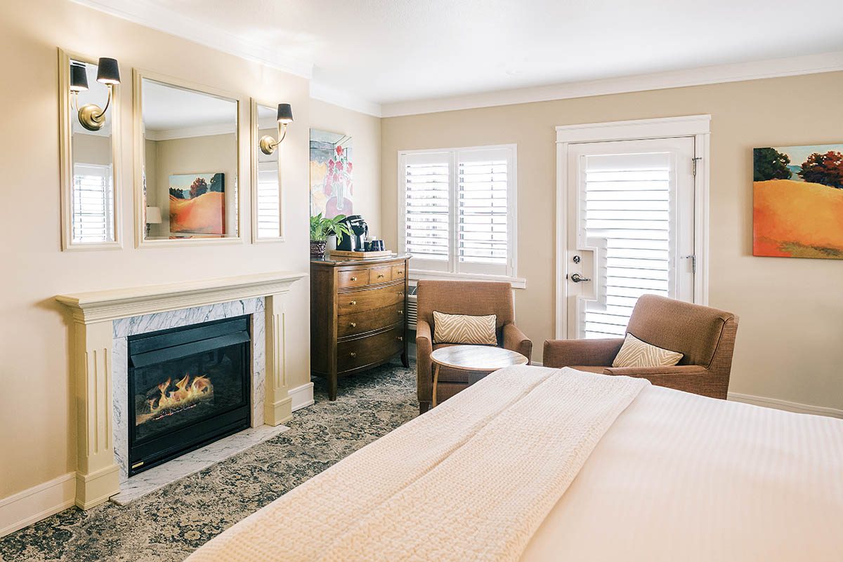 Plaza Deluxe king room at Napa River Inn with a marble-surround gas fireplace, framed wine-country artwork, plantation-shuttered French doors, and twin armchairs.