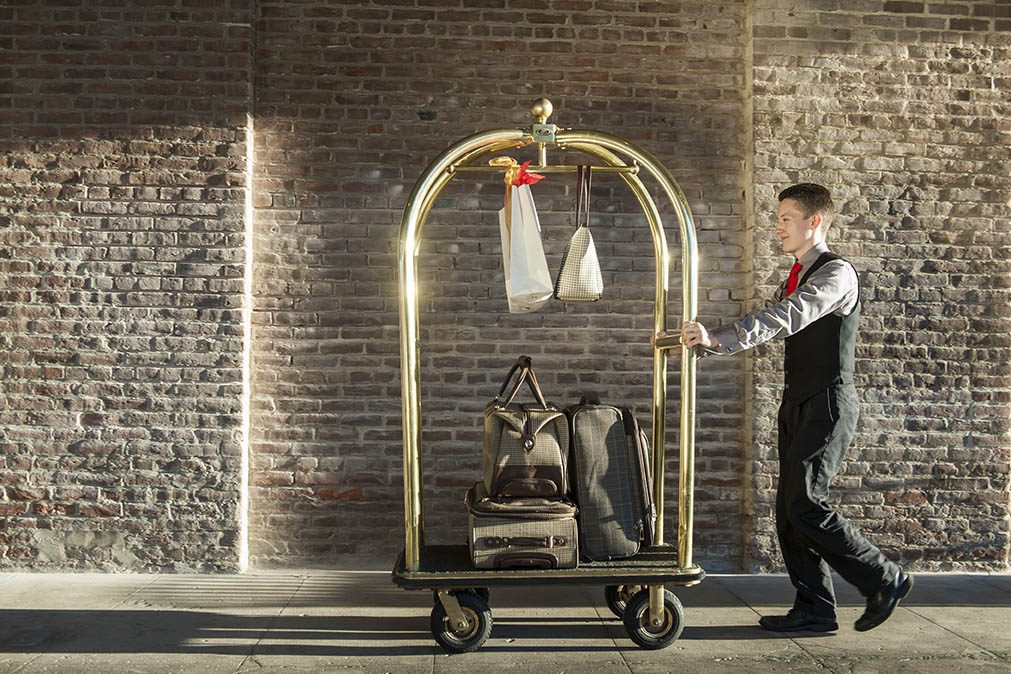 A Napa River Inn bellman in vest and tie pushing a brass bellhop cart loaded with luggage along the historic brick exterior of the Napa Mill.