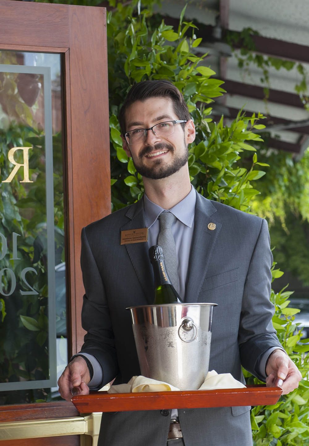 Man in a suit holding a tray with a wine bottle in an ice bucket, standing by greenery and a window.
