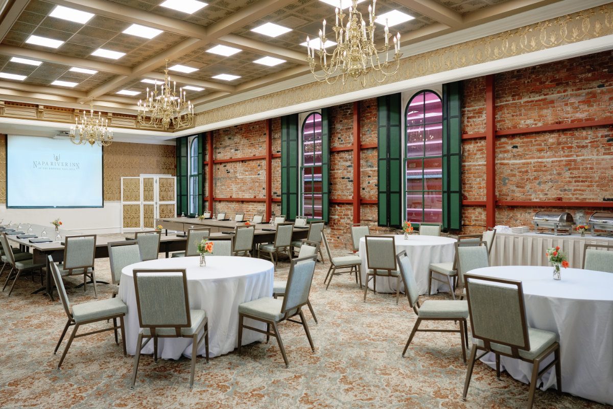 Hatt Hall event space at Napa River Inn set with round banquet tables and a U-shape boardroom, featuring exposed brick walls, arched windows, and crystal chandeliers inside the Historic Napa Mill.