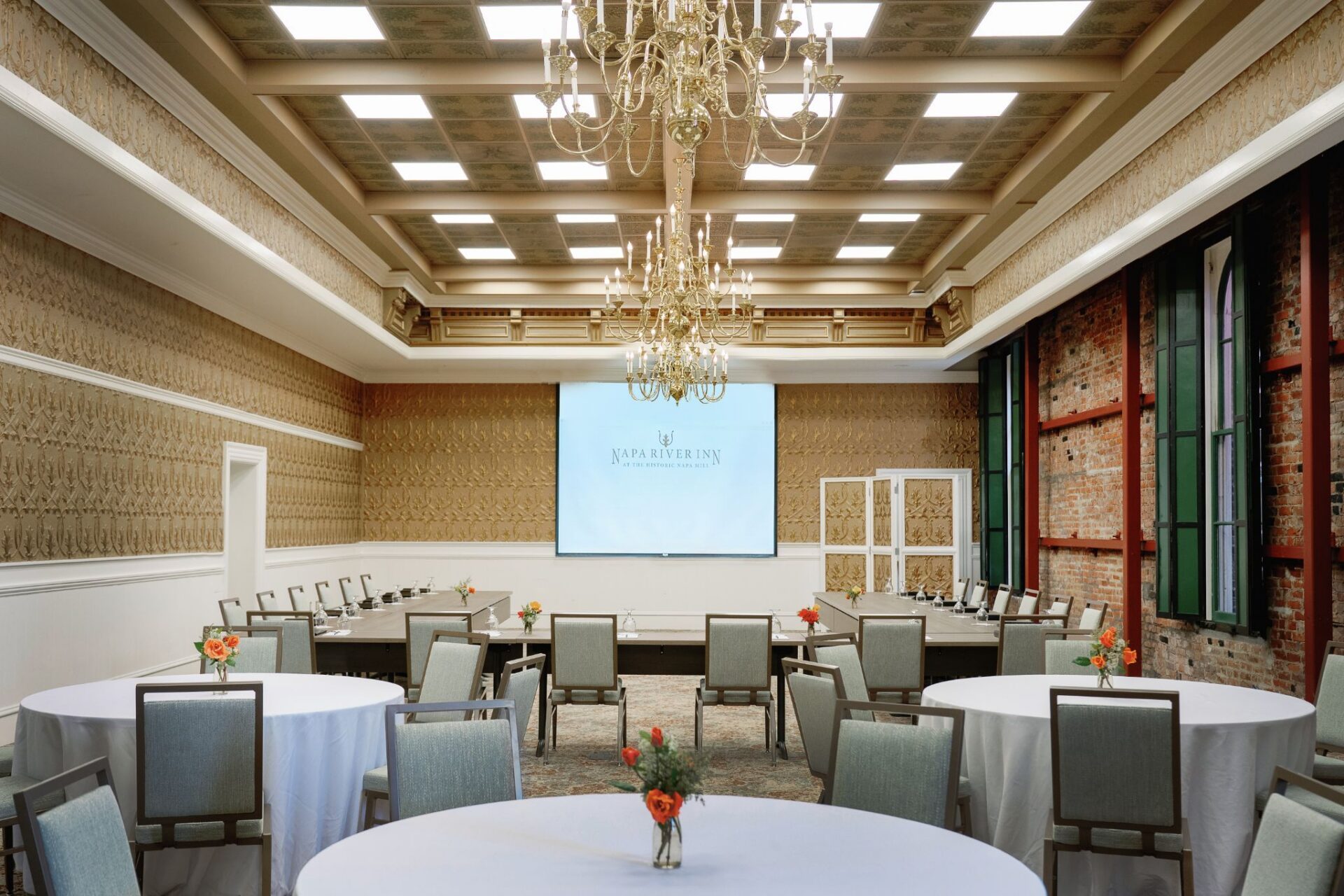 Hatt Hall meeting room at Napa River Inn arranged for a hybrid event with a projection screen, U-shape conference seating, and grand chandeliers under a coffered ceiling.