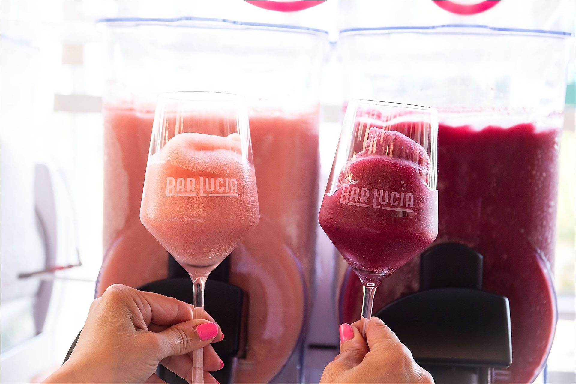 Two Bar Lucia wine glasses filled with strawberry and red-wine frosé held in front of frozen slushie machines in Downtown Napa.