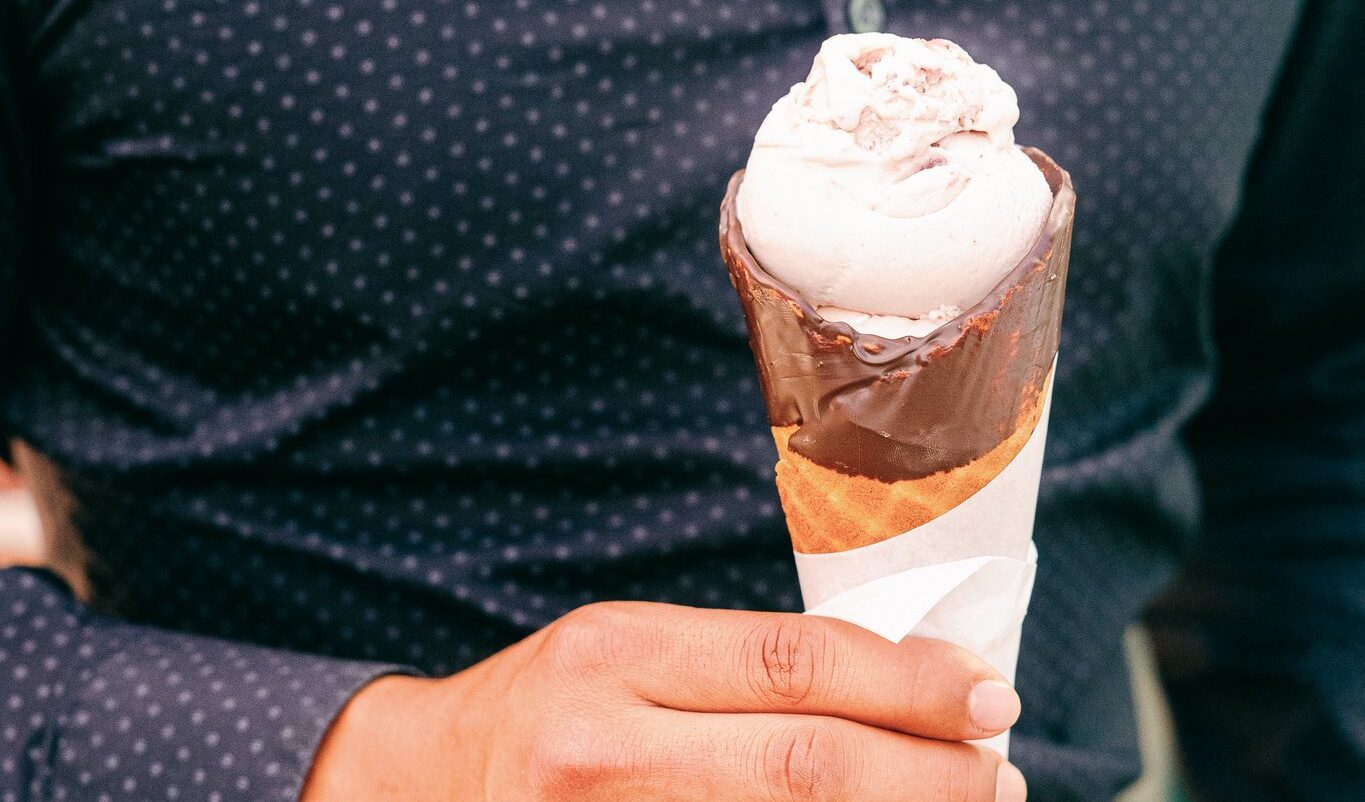 A hand holding a scoop of strawberry ice cream in a chocolate-dipped waffle cone wrapped in paper, from an ice cream shop in Downtown Napa.
