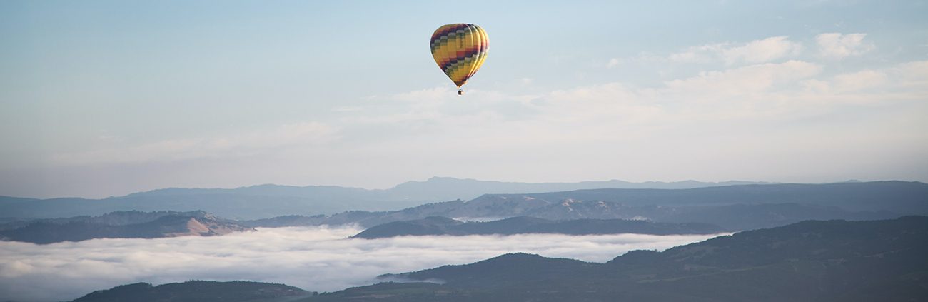 A colorful hot air balloon drifting above fog-filled Napa Valley vineyards and rolling hills at sunrise.